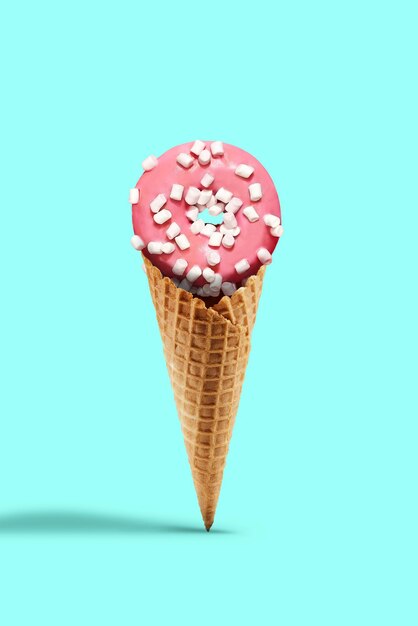 Ciambella dolce glassata da glassa rosa cosparsa di marshmallow in un cono di cialda su sfondo turchese. Concetto di cibo, dolcetti e alimentazione malsana. Primo piano, copia spazio