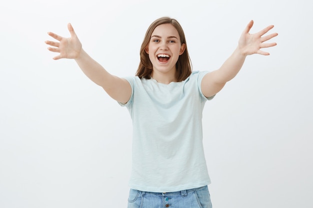 Ciao da molto tempo che non ci vediamo. Amichevole gioiosa bella ragazza con i capelli castani corti tirando le palme in segno di saluto verso il dire ciao dare un caloroso benvenuto e abbraccio amico sul muro grigio sorridendo felicemente