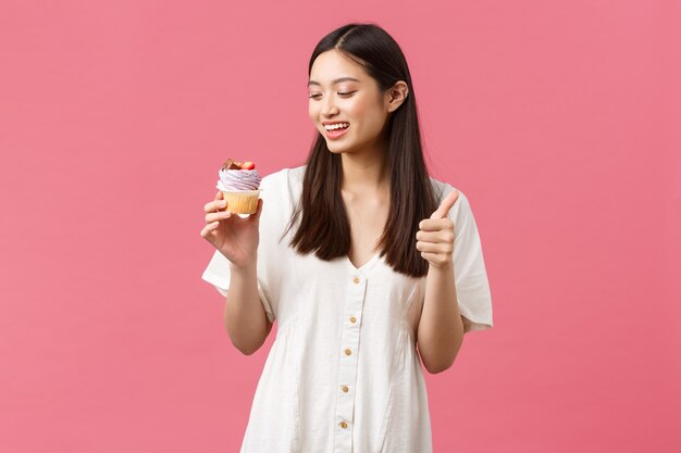 Cibo, caffè e ristoranti, concetto di stile di vita estivo. Una cliente femminile sorridente e felice consiglia deliziosi cupcake allo spettacolo di prodotti da forno, mostrando il pollice in su e guardando il dessert con desiderio di mordere