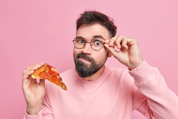 Cibo delizioso nocivo. L'uomo barbuto bello ha uno spuntino gustoso tiene la mano sul bordo degli occhiali tiene una fetta di appetitosa pizza italiana ha abitudini alimentari dannose e malsane