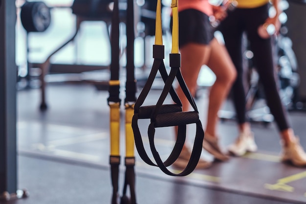 Cinghie di sospensione sportive in primo piano e due ragazze fitness che parlano in sottofondo nella moderna palestra.