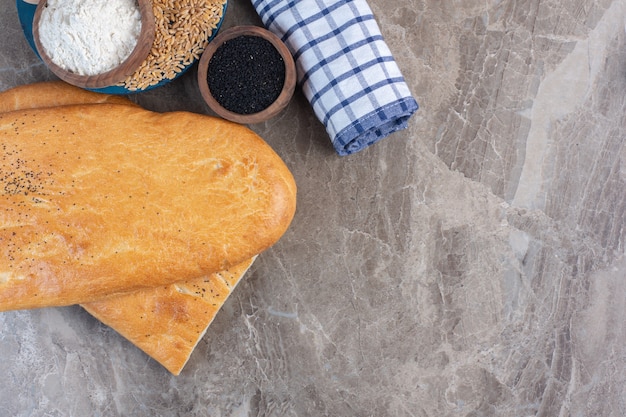 Ciotola di farina e mucchio di grano su un vassoio accanto alla ciotola di sesamo nero, rotolo di asciugamano e pagnotte di pane su marmo.