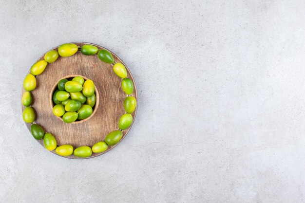 Ciotola di kumquat circondata da un cerchio di kumquat su una tavola di legno su fondo marmo.