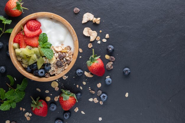 Ciotola di muesli d'avena con yogurt, mirtilli freschi, fragole, kiwi menta e tavola di noci per una sana colazione, vista dall'alto, spazio copia, piatto Concetto di menu per la colazione sana. Sulla roccia nera