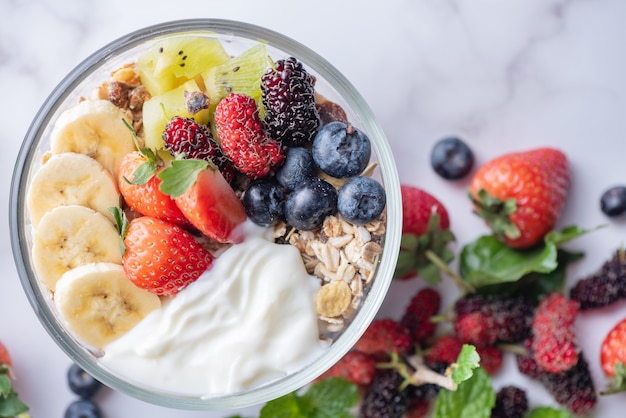 Ciotola di muesli d'avena con yogurt, mirtilli freschi, gelso, fragole, kiwi, banana, menta e frutta secca per una sana colazione, vista dall'alto, spazio copia, piatto. concetto di cibo vegetariano.