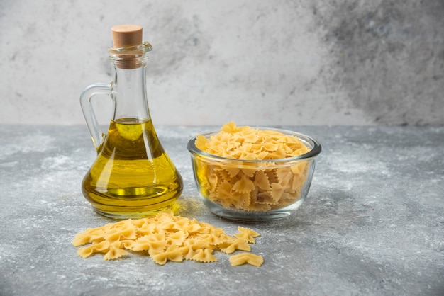 Ciotola di pasta cruda farfalle con bottiglia di olio d'oliva su sfondo marmo.