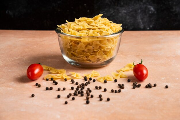 Ciotola di pasta cruda, grani di pepe e pomodori freschi sulla tavola arancione.