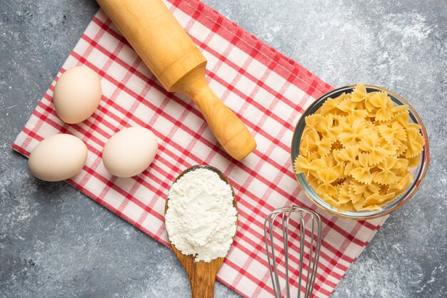 Ciotola di pasta cruda, uova, cucchiaio di farina e mattarello sul tavolo di marmo con tovaglia.