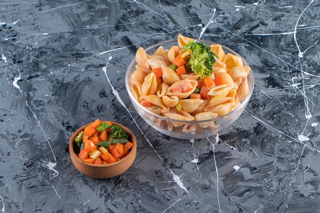 Ciotola di vetro di gustosa pasta di conchiglie con insalata fresca su superficie di marmo.