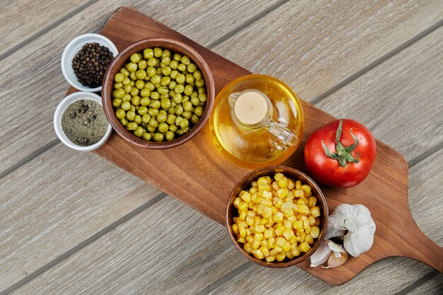 Ciotole di mais dolce bollito e piselli, spezie, olio e verdure su una tavola di legno.