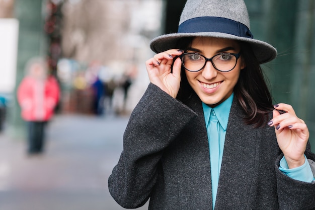 Città ritratto elegante affascinante giovane donna in cappotto grigio, cappello che cammina sulla strada. Occhiali neri moderni, sorridenti, che esprimono vere emozioni felici, stile di vita di lusso, modello alla moda.