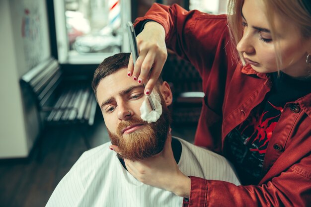 Cliente durante la rasatura della barba nel negozio di barbiere. Barbiere femminile al salone. Parità dei sessi. Donna nella professione maschile.