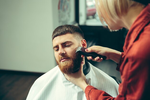 Cliente durante la rasatura della barba nel negozio di barbiere. Barbiere femminile al salone. Parità dei sessi. Donna nella professione maschile.