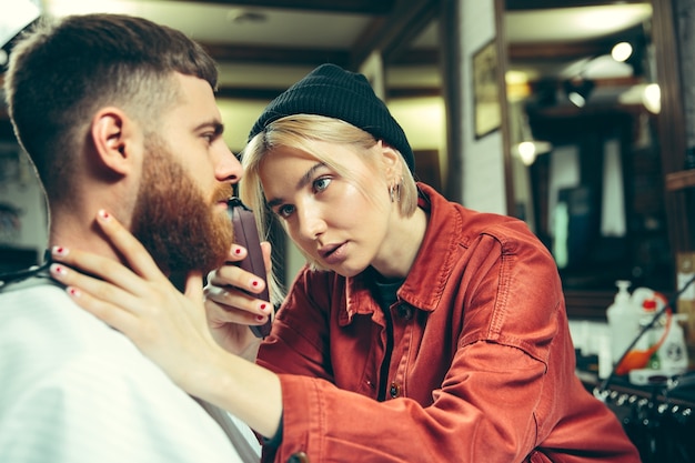 Cliente durante la rasatura della barba nel negozio di barbiere. Barbiere femminile al salone. Parità dei sessi