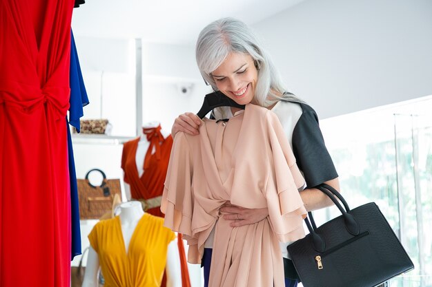 Cliente femminile allegro che gode dello shopping, applicando il vestito con il gancio. Donna che sceglie i vestiti nel negozio di moda. Shopping o concetto di vendita al dettaglio