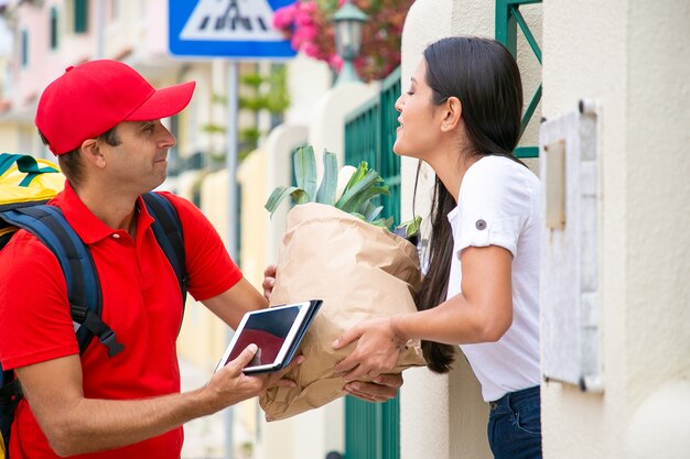 Cliente femminile felice che riceve cibo dal negozio di alimentari, prendendo il pacchetto dal corriere al suo cancello. Concetto di servizio di spedizione o consegna