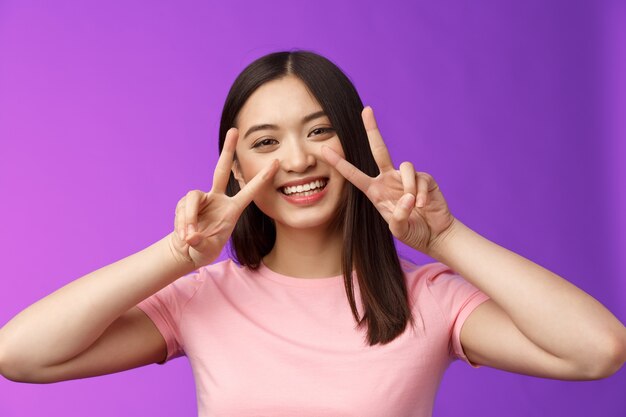 Close-up amichevole positiva in uscita bella ragazza asiatica mostra pace, segni di vittoria caro friendhsip rimanere ottimista, sorridente ampiamente, avendo divertenti vacanze estive, stand sfondo viola.