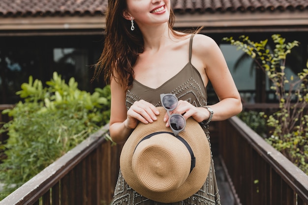 Close up dettagli mani che tengono cappello di paglia e occhiali da sole, accessori eleganti, giovane donna attraente in abito elegante, stile estivo, tendenza moda, vacanze, posa su villa tropicale, sorridente, felice