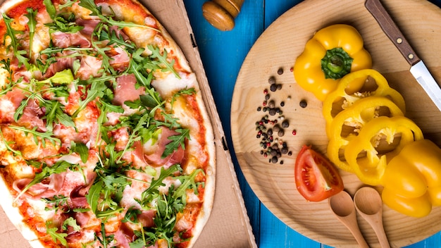 Close-up di pizza con pancetta e foglie di rucola vicino a peperone a fette; pomodoro; aglio e spezie sul tavolo