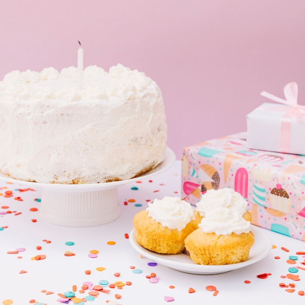 Close-up di scatole regalo avvolto; torta alla vaniglia e cupcakes con coriandoli colorati sulla scrivania bianca