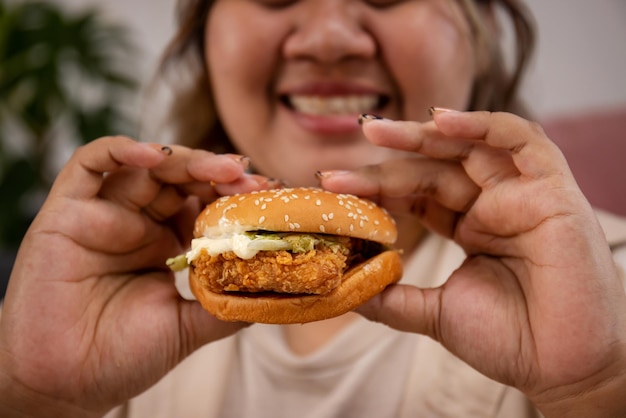 Close up felice donna grassa asiatica godere di mangiare delizioso hamburger sul soggiorno