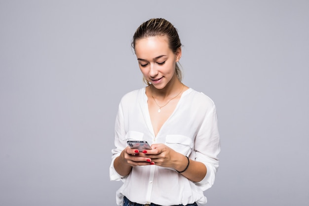 Close up foto di attraente ragazza sorridente in piedi contro il muro grigio e digitando sms sul suo smartphone