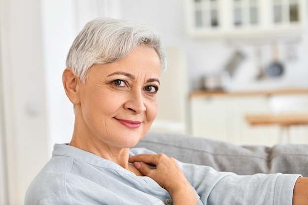 Close up foto interna di elegante donna anziana titolare di pensione o di rendita in un momento di relax a casa comodamente seduto sul divano dopo la doccia, indossando un accogliente accappatoio di cotone con grazioso sorriso affascinante