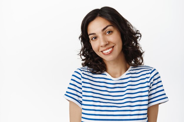 Close up ritratto di attraente ragazza bruna con i capelli ricci, sorridente denti bianchi, guardando carino e sciocco alla telecamera, in piedi felice su sfondo bianco.