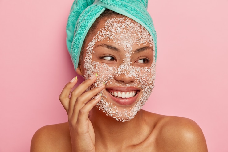 Foto gratuito close up ritratto di donna felice massaggia le guance, applica lo scrub al sale marino, distoglie lo sguardo, ha un sorriso gentile, mostra i denti bianchi, indossa un asciugamano turchese, felice di ricevere trattamenti termali.