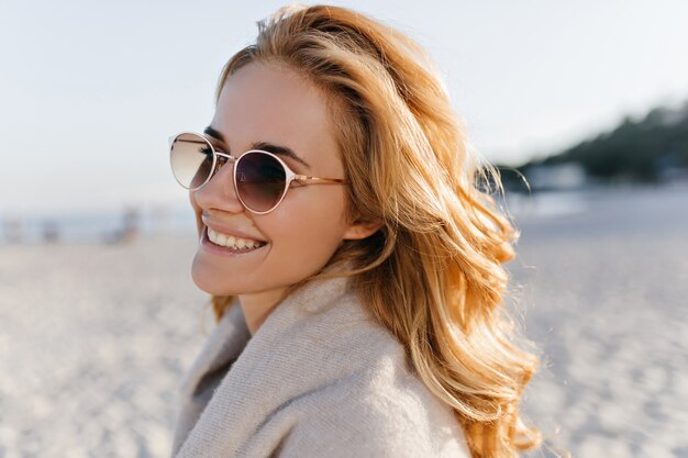 Close-up ritratto di donna positiva con capelli ondulati blinde vestita in maglione di cashmere beige e occhiali da sole in spiaggia.