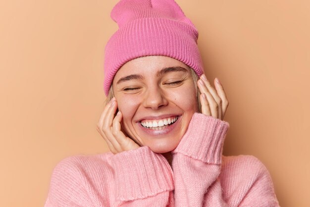 Close up ritratto di felice bella donna tiene le mani sul viso sorride volentieri tiene gli occhi chiusi mostra denti bianchi essendo di buon umore indossa cappello rosa e maglione isolato sopra la parete beige dello studio