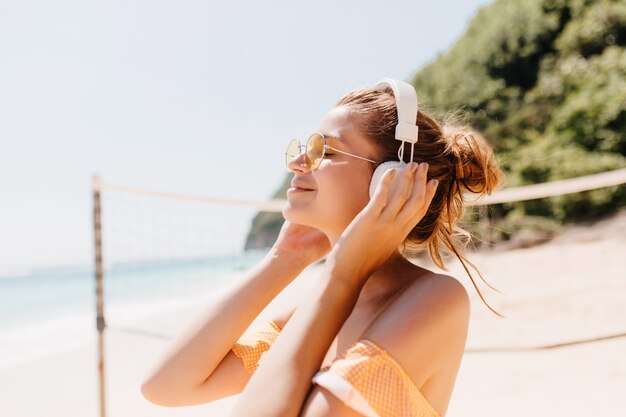 Close-up ritratto di gioiosa donna abbronzata rilassante con la musica preferita in spiaggia. Colpo esterno del modello femminile sorridente in cuffie che trascorrono del tempo al resort.