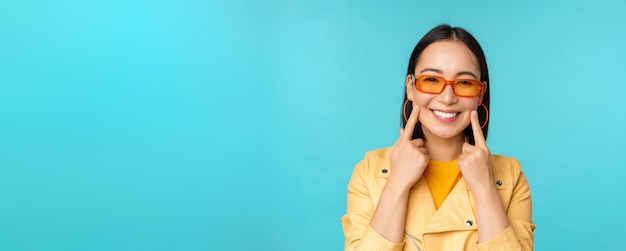 Close up ritratto di giovane donna asiatica in occhiali da sole sorridente e guardando romantico in piedi felice su sfondo blu