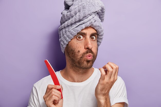 Close up ritratto di giovane ragazzo con la barba lunga soffia sulle unghie, fa manicure a casa, tiene la lima per unghie, cura della sua bellezza e igiene, indossa telo da bagno e maglietta bianca, isolato sul muro viola.