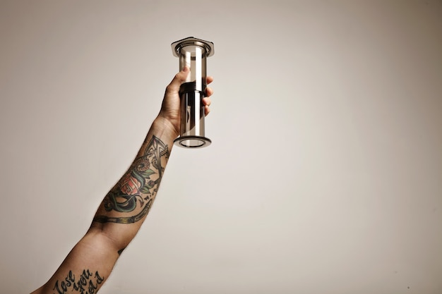 Close up ritratto di un tatuato di mano dell'uomo che tiene chiaro grigio chiaro aeropress non tradizionali attrezzature per la preparazione del caffè in alto nell'aria isolato su bianco