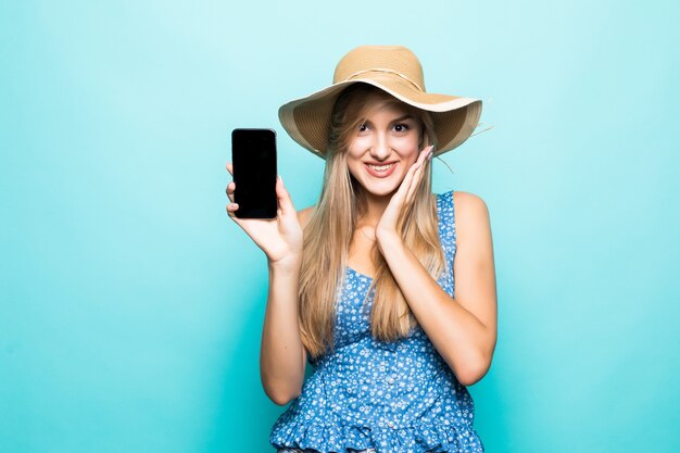 Close up ritratto di una giovane donna allegra in abito e cappello estivo che mostra il telefono cellulare con schermo vuoto isolato su sfondo blu