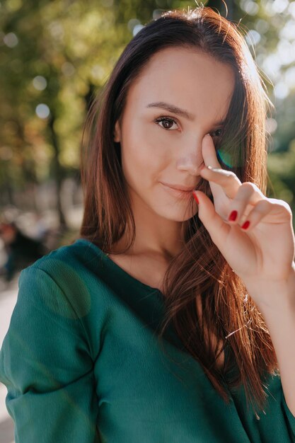 Close up ritratto esterno di elegante bella ragazza sta guardando la fotocamera e coprendo il viso mentre si cammina nel parco soleggiato con alberi verdi in una calda giornata estiva