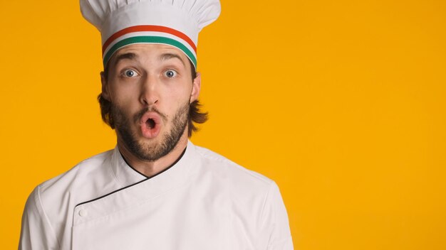 Close up sorpreso chef maschio vestito in uniforme guardando stupito la fotocamera su sfondo colorato Uomo barbuto in cappello da chef dimenticato di acquistare alcuni prodotti per il lavoro in cucina