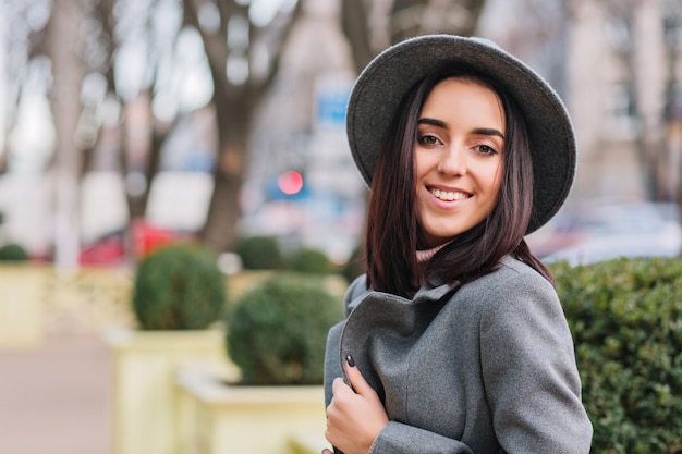 Closeup ritratto alla moda giovane donna in cappello grigio, cappotto che cammina sulla strada nel parco cittadino. Capelli castani, sorriso, umore allegro, aspetto elegante.