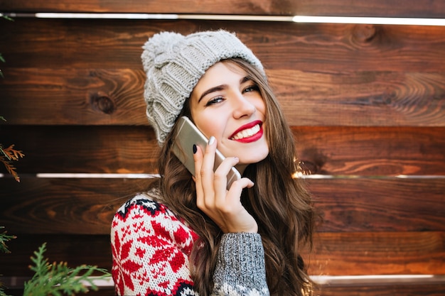 Closeup ritratto bella ragazza con i capelli lunghi e le labbra rosse su legno. Indossa un caldo cappello e un maglione invernali, parla al telefono e sorride.