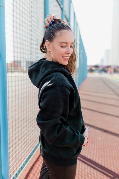 Closeup ritratto di bella ragazza con i capelli lunghi sorridente con gli occhi chiusi sullo sfondo del campo sportivo Indossa jeans e pullover neri Vista dal retro