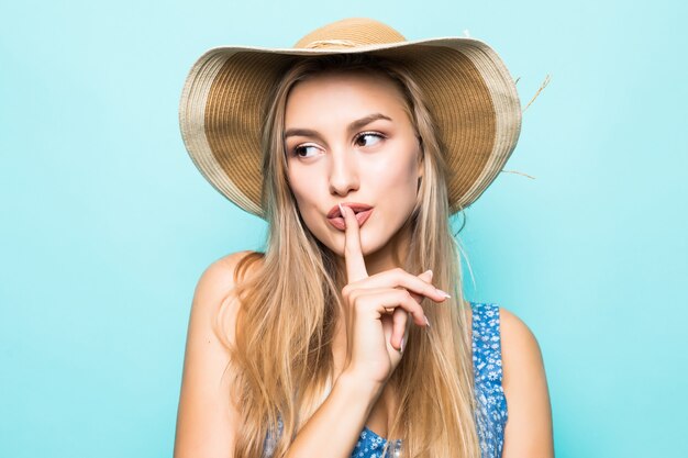 Closeup ritratto di donna affascinante europea che indossa un grande cappello di paglia che mostra il dito sulle labbra per mantenere il segreto isolato su sfondo blu