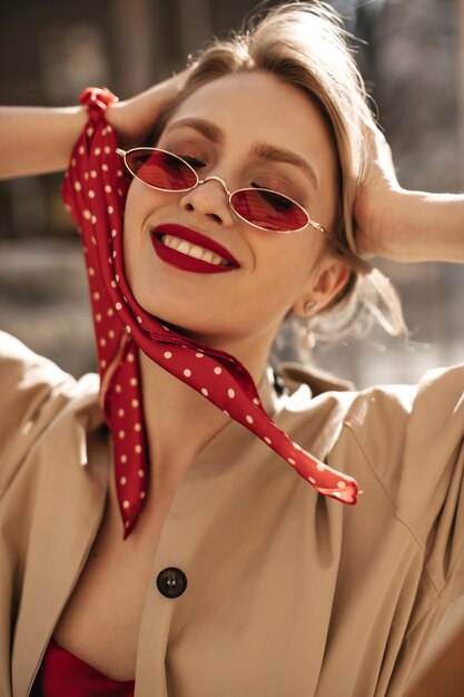 Closeup ritratto di donna attraente con trucco splendido sorridente fuori Donna bionda in occhiali da sole rossi e trench beige tocca i capelli