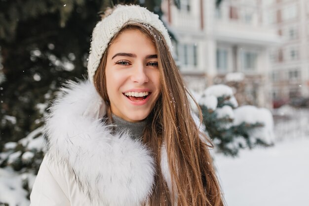 Closeup ritratto di gioiosa donna sorridente in cappello lavorato a maglia in posa all'aperto sulla strada piena di neve. Signora bionda allegra con gli occhi azzurri che gode del periodo invernale che trascorre il fine settimana nel cortile.