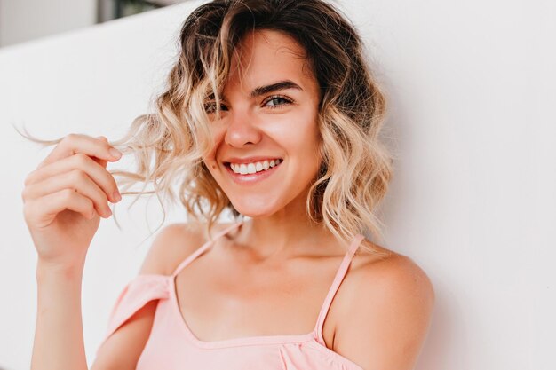 Closeup ritratto di incantevole ragazza ridente che gioca con i capelli ondulati Foto di donna abbronzata interessata con un bel sorriso
