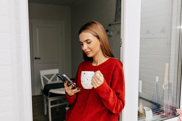 Closeup ritratto di splendida giovane donna bionda che indossa un maglione rosso in piedi accanto alla finestra È felice sorridente smartphone a scorrimento