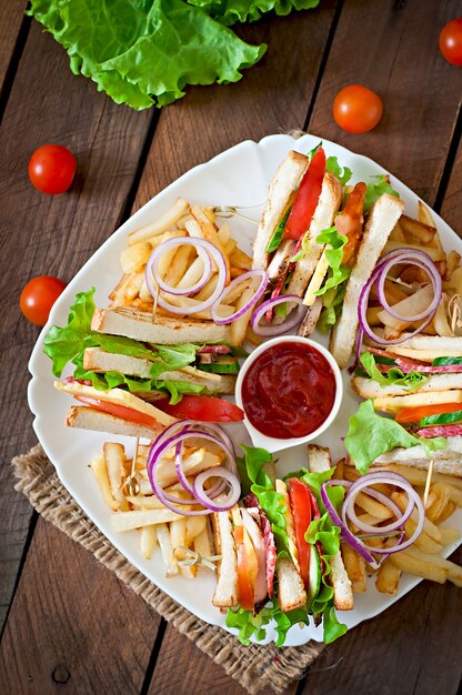 Club sandwich con formaggio, cetriolo, pomodoro, carne affumicata e salame. Servito con patatine fritte.