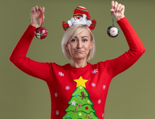 Clueless donna bionda di mezza età che indossa la fascia di Babbo Natale e maglione di Natale sollevando palline di Natale guardando la telecamera isolata su sfondo verde oliva