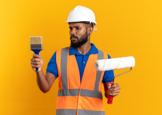 Clueless giovane afro-americano costruttore uomo in uniforme con casco di sicurezza tenendo il rullo di vernice e guardando il pennello isolato su sfondo arancione con spazio di copia
