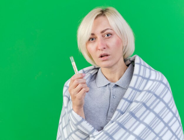 Clueless giovane bionda malata donna slava avvolta in plaid tiene termometro isolato sulla parete verde con copia spazio